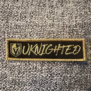 Peut inclure: Un écusson brodé noir et or avec le texte "UKNighted" en lettres dorées. L'écusson présente un logo stylisé de casque de chevalier.