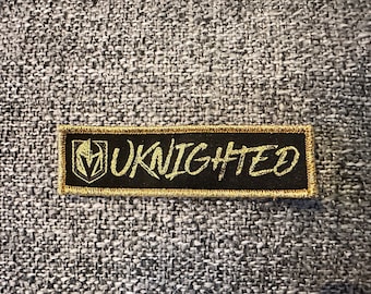 Puede incluir: Un parche bordado negro y dorado con el texto "UKNighted" en letras doradas. El parche presenta un logotipo estilizado de casco de caballero.