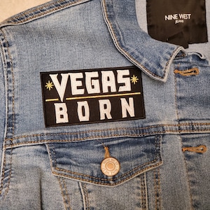 May include: A blue denim jacket with a black patch that says "*VEGAS* BORN" in white lettering with a gold line above the word "BORN".