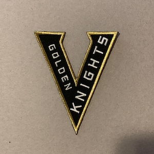 May include: Black and gold embroidered patch with a white "V" in the centre. The text "Golden Knights" is written vertically on either side of the "V".