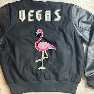 Può includere: Giacca da baseball nera con maniche in pelle. La parte posteriore della giacca ha un "VEGAS" bianco ricamato sul retro e una toppa di fenicottero rosa.