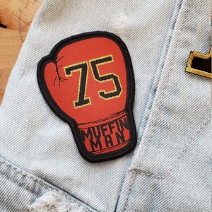 Può includere: Un cerotto a forma di guanto da boxe rosso con il numero 75 in lettere dorate e le parole "Muffin Man" in lettere nere.