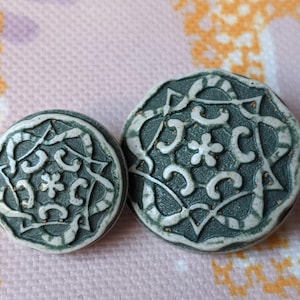 Könnte beinhalten: Zwei dekorative Knöpfe mit einem verzierten Design. Die Knöpfe sind dunkelgrau mit einem weißen und goldenen Blumenmuster. Die Knöpfe sind unterschiedlich groß, der größere Knopf befindet sich rechts.