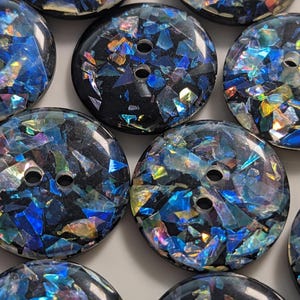 Könnte beinhalten: Nahaufnahme mehrerer runder Knöpfe mit jeweils zwei Löchern. Die Knöpfe haben eine schwarze Basis mit schillernden blauen, grünen und goldenen Fragmenten, die in die Oberfläche eingebettet sind. Die Knöpfe sind in einem engen Muster angeordnet.
