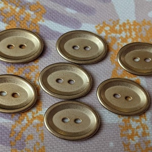 May include: Six gold oval-shaped buttons with two holes each. The buttons have a raised edge and a smooth, shiny surface.