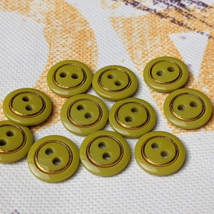 Op de afbeelding: Een verzameling van 11 ronde, groene knopen met een gouden rand. De knopen hebben elk twee gaten.