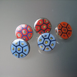 May include: Five round, decorative buttons with a silver rim. The button faces feature a colorful geometric design with red, blue, and orange backgrounds, and yellow and blue starburst patterns. The buttons are arranged on a reflective surface.