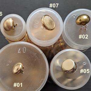 Könnte beinhalten: Nahaufnahme von zehn goldfarbenen Knöpfen in durchsichtigen Plastikbehältern. Die Knöpfe sind mit den Nummern 01 bis 07 beschriftet.