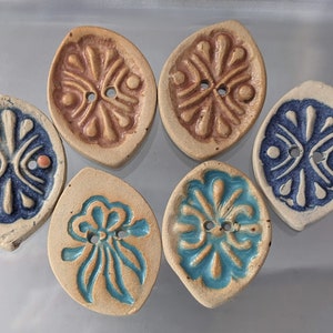 Könnte beinhalten: Sechs Keramikknöpfe in verschiedenen Farben und Designs. Die Knöpfe sind blattförmig und haben ein Loch in der Mitte. Die Designs umfassen ein blaues und weißes Blumenmuster, ein braunes und weißes Blumenmuster und ein blaues und weißes geometrisches Muster.