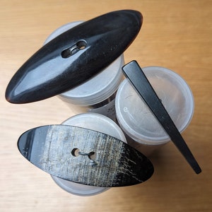 Puede incluir: Tres botones de cuerno negro, dos ovalados y uno redondo, con un agujero en el centro de cada uno. Los botones se muestran sobre recipientes de plástico transparente.