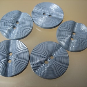May include: Five pale blue, circular buttons with a spiral groove design. Each button has two holes for sewing. The buttons are made of a smooth, glossy material and are arranged in a scattered pattern.