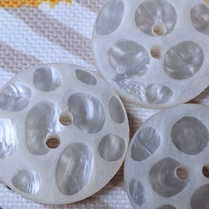 Op de afbeelding: Close-up van verschillende crèmekleurige, ronde knopen met een parelmoerglans. Elke knoop heeft een reeks onregelmatige, lichtgekleurde gaten en een centraal gat om te naaien. De knopen zijn gerangschikt op een gestreepte stof.