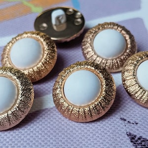 May include: Close-up of six decorative buttons. Each button has a white centre surrounded by a textured, gold-coloured rim. One button has a gold-coloured shank on the back. The buttons are arranged on a light purple and white patterned surface.