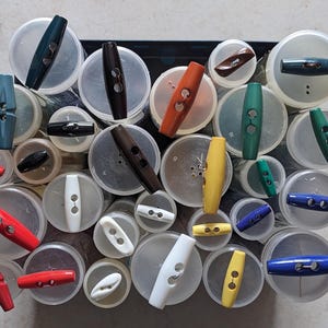 Könnte beinhalten: Sortiment an bunten Kunststoff-Toggle-Knöpfen in verschiedenen Größen und Formen. Die Knöpfe sind kreisförmig angeordnet und in durchsichtigen Plastikbehältern ausgestellt.