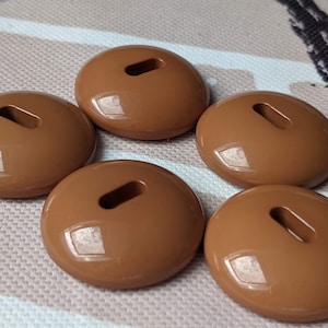 Peut inclure: Cinq boutons ovales bruns brillants. Chaque bouton a une fente rectangulaire au centre. Les boutons sont disposés de manière légèrement éparpillée.