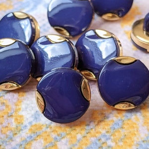Może przedstawiać: Kolekcja granatowych guzików ze złotymi akcentami. Guziki są okrągłe, z błyszczącym wykończeniem i trzpieniem z tyłu. Złote akcenty znajdują się po bokach guzików.