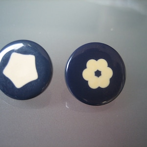 May include: Two round, blue ceramic buttons with white designs. One button has a five-pointed star design and the other has a flower design.