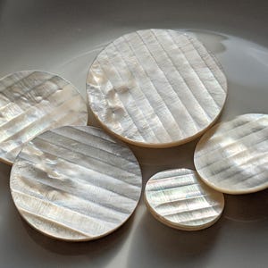 Op de afbeelding: Vijf ronde, iriserende onderzetters van parelmoer met een geribbelde textuur. De onderzetters variëren in grootte en zijn gerangschikt op een wit oppervlak. Het parelmoer heeft een parelmoerglans die licht reflecteert.