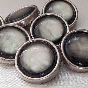May include: A set of seven round, silver-toned buttons with a black rim and a white, pearlescent centre. The buttons have a smooth, shiny finish.