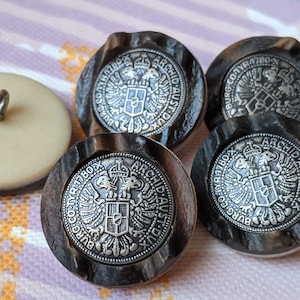 May include: A collection of vintage buttons. One button is off-white with a silver shank, while the others are dark brown with a silver emblem featuring an eagle and text. The buttons have a unique, antique aesthetic.