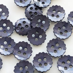 Op de afbeelding: Een verzameling marineblauwe knopen met geschulpte randen en vier gaten. De knopen zijn verspreid over een wit oppervlak, wat hun glanzende afwerking en uniforme ontwerp benadrukt. Ideaal voor naai- en knutselprojecten.
