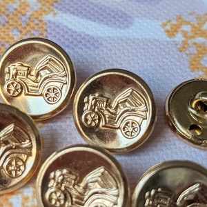 May include: Six gold-toned metal buttons featuring a raised design of a vintage car. The buttons have a smooth, shiny finish.