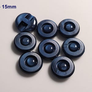 Puede incluir: Ocho botones azul marino con acabado brillante. Cada botón tiene un diseño circular con un centro elevado y un diámetro de 1,5 cm. Un botón tiene un diseño en forma de cruz. El texto "#03 - 15mm" está en la esquina superior izquierda.