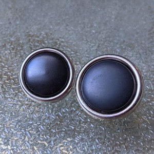 Könnte beinhalten: Zwei schwarze Knöpfe mit silbernen Rändern. Die Knöpfe sind rund und haben eine glatte, glänzende Oberfläche.