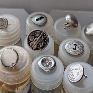 Op de afbeelding: Assortiment van zilveren en witte knopen in verschillende ontwerpen, waaronder een muzieknootontwerp, een bloemenontwerp en een effen rond ontwerp. De knopen worden gepresenteerd in doorzichtige plastic containers.