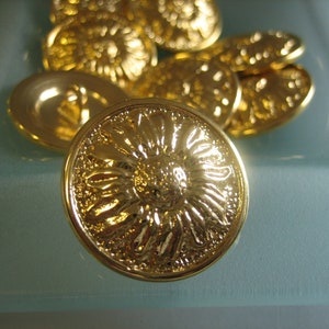 Könnte beinhalten: Eine Gruppe goldfarbener Metallknöpfe mit einem dekorativen Blumendesign. Die Knöpfe liegen auf einer hellblauen Oberfläche.