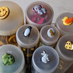 Könnte beinhalten: Acht durchsichtige Plastikbehälter mit Deckeln, die jeweils eine Vielzahl von bunten Knöpfen enthalten. Die Deckel sind mit verschiedenen Tier- und Smiley-Motiven verziert. Die Knöpfe sind in verschiedenen Formen, Größen und Farben.