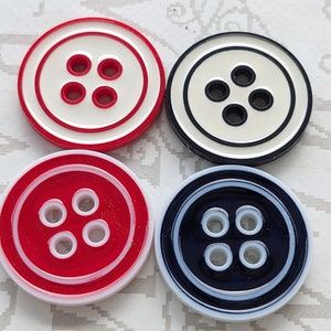 Könnte beinhalten: Vier runde Knöpfe in Rot, Weiß und Marineblau. Jeder Knopf hat einen kontrastierenden Rand und vier Löcher. Die Knöpfe sind ein klassisches Design, geeignet zum Nähen oder Basteln.
