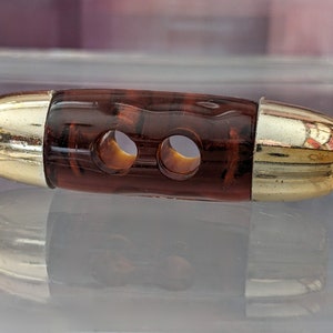 Pode incluir: Um botão decorativo com um centro castanho escuro marmoreado e extremidades em metal dourado. O botão tem dois orifícios e uma forma ligeiramente curva, adequado para projetos de costura ou artesanato.