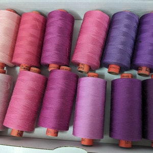 Op de afbeelding: Een doos met 12 spoelen garen in tinten roze en paars. De spoelen zijn gerangschikt in rijen van drie, waarbij de kleuren geleidelijk overgaan van lichtroze naar donkerpaars.