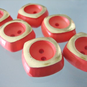 Puede incluir: Primer plano de seis botones cuadrados. Los botones son de un color coral vibrante con un borde dorado en el borde superior. Cada botón tiene dos pequeños agujeros para coser. Los botones están dispuestos en un patrón ligeramente disperso.