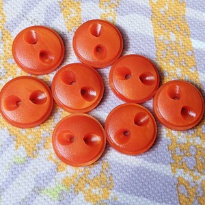 Könnte beinhalten: Acht orangefarbene Knöpfe mit zwei Löchern. Die Knöpfe sind rund und haben eine leicht gewölbte Oberfläche. Sie bestehen aus einem glatten Material und liegen auf einem gemusterten Stoff. Ideal für Nähprojekte.