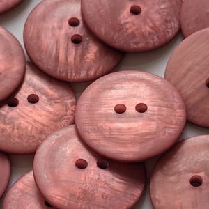 Op de afbeelding: Close-up van verschillende grote, ronde, roze knopen. Elke knoop heeft twee of drie gaten om op te naaien. De knopen hebben een licht iriserende afwerking, waardoor ze een subtiele glans hebben. De knopen zijn verspreid.