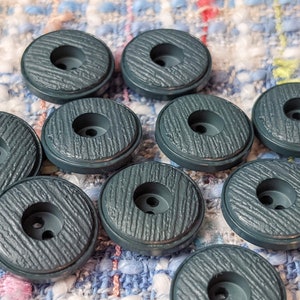 Könnte beinhalten: Eine Sammlung von zehn runden, teal-farbenen Knöpfen. Jeder Knopf hat eine strukturierte, holzmaserungsähnliche Oberfläche und ein zentrales Loch mit zwei kleineren Löchern darunter. Die Knöpfe sind auf einem mehrfarbigen, strukturierten Stoff angeordnet.