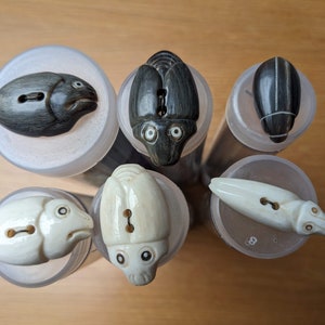 Pode incluir: Uma coleção de seis botões esculpidos, cada um representando uma criatura diferente. Os botões são em tons de preto e creme, com esculturas detalhadas de peixes, insetos e outros animais. São apresentados em tubos de plástico transparente.