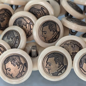 May include: A collection of off-white buttons with a raised design of a person's face in a profile view. The buttons have a smooth, slightly textured surface.