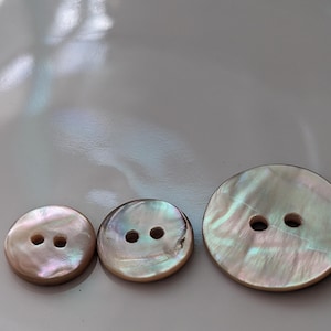 May include: Three iridescent mother-of-pearl buttons of varying sizes. The buttons have two holes each and display a range of colors, including pink, white, and green. The largest button is approximately 1.25 inches in diameter.