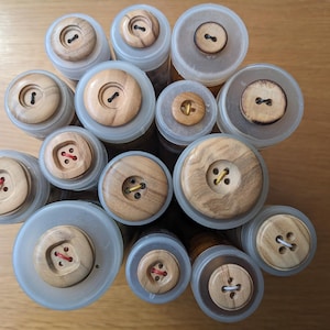 Op de afbeelding: Een verzameling houten knopen in verschillende maten en kleuren, gepresenteerd in doorzichtige plastic containers. De knopen hebben een natuurlijke houten afwerking en zijn in een cirkelvormige opstelling gerangschikt.