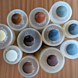 May include: A collection of ten small round containers with lids, each containing a different colour of paint. The colours include brown, white, dark brown, light brown, red, dark blue, light blue, and grey.
