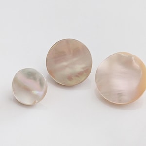 Puede incluir: Tres botones circulares hechos de nácar iridiscente. Los botones presentan una superficie lisa y pulida con variaciones naturales nacaradas en tonos rosa suave, crema y blanco, mostrando un brillo sutil y una textura orgánica.
