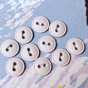 Peut inclure: Dix boutons ronds beiges avec deux trous chacun, disposés sur un tissu à motifs. Les boutons sont espacés uniformément et semblent être en plastique ou dans un matériau similaire. Le tissu de fond présente un motif floral bleu clair et blanc.