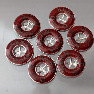 Op de afbeelding: Zeven rode metalen knopen met een zilveren midden en het woord "KERRY OTTO" rond de rand.