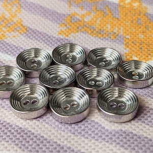 Peut inclure: Dix boutons en métal argenté avec un motif de cercles concentriques. Les boutons sont disposés en cercle sur un fond de tissu rayé.