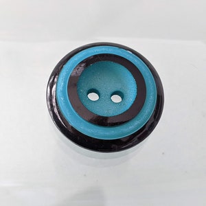 Op de afbeelding: Een ronde knoop met twee gaten en een gelaagd ontwerp met een glanzende zwarte buitenrand, een heldere turquoise middelste ring en een getextureerd turquoise midden. De knoop heeft een gladde, gepolijste afwerking op de zwarte delen.