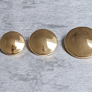 Op de afbeelding: Drie gouden metalen knopen in verschillende maten. De knopen zijn rond en hebben een gladde, glanzende afwerking.