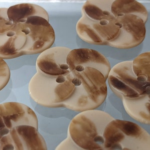 May include: A group of eight brown and beige buttons with a flower-like design. The buttons have a swirl pattern and three holes.